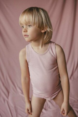 Chłopiec o włosach blond stoi w koszulce na ramiączka tank top i majtkach chłopięcych w kolorze śmietankowego różu na tle z tej samej różowej dzianiny. Chłopiec patrzy w bok